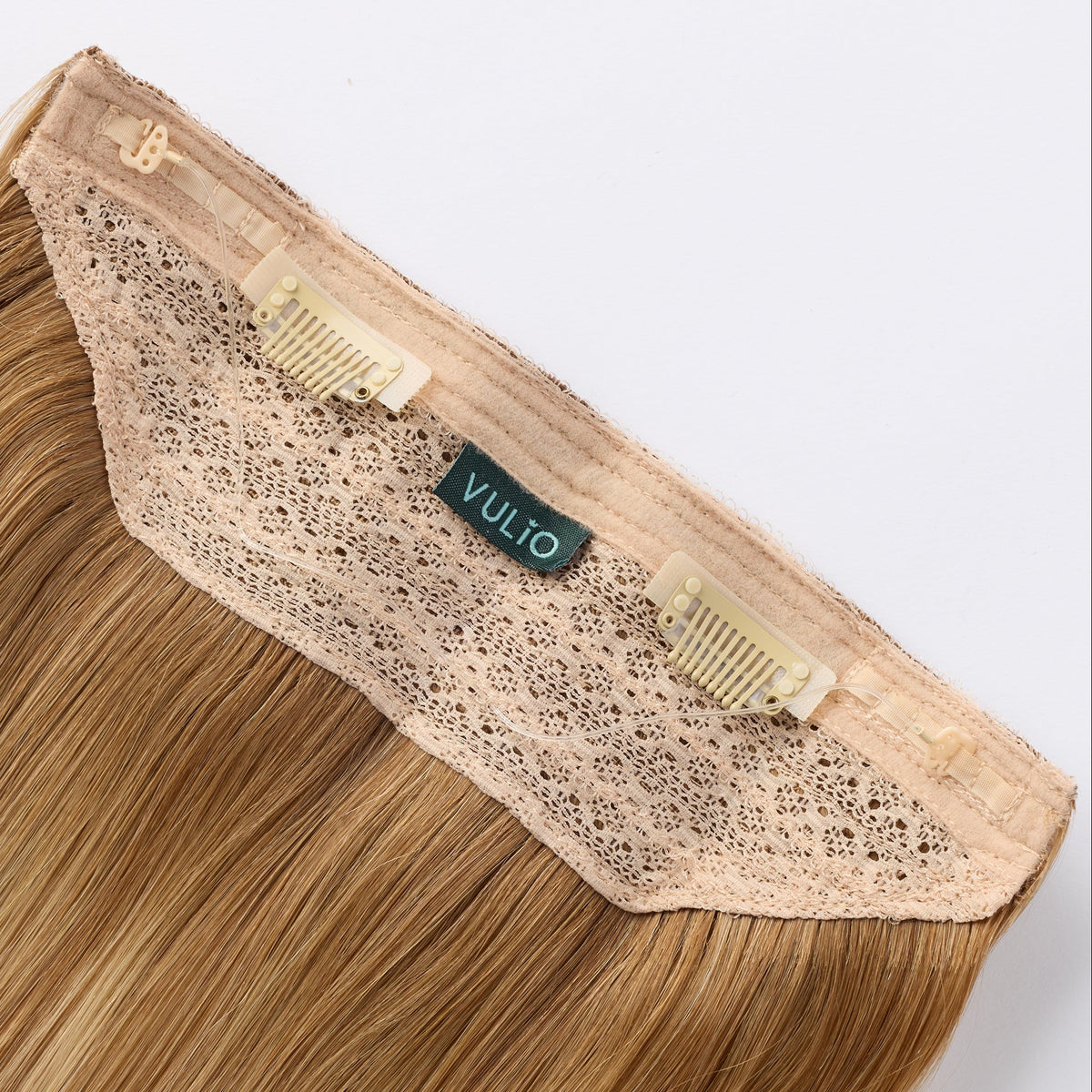 A close-up of a VULIO clip-in hair extension showing the lace front, adhesive clips, and fine hair strands in a warm blonde shade against a light gray background.